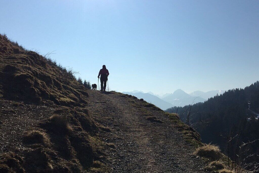 Eine Person geht auf einem Bergpfad, der von einem klaren Himmel und fernen Bergen gesäumt wird.