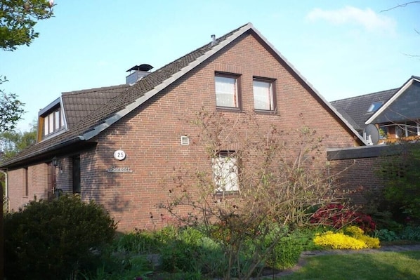 A two-story brick house with a gabled roof, a small front porch, and a garden with various plants and flowers. - Stender "An der olen Au" (Winsen)