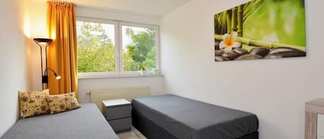 A bedroom with two single beds, a bedside table, a lamp, a window with curtains, and a wall-mounted picture.