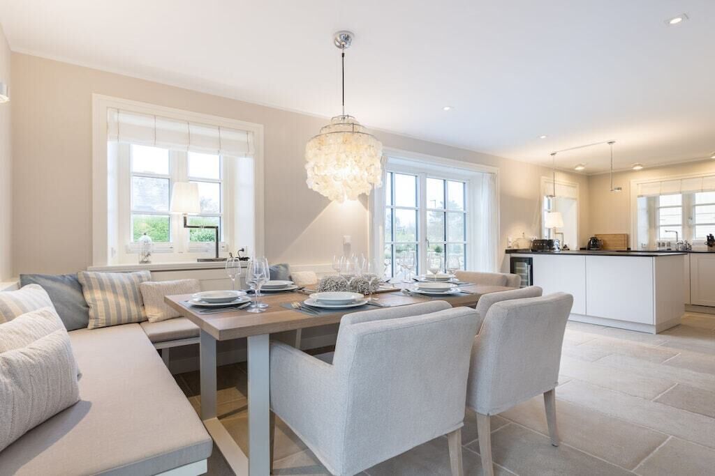 A modern kitchen with a dining area, featuring a chandelier, large windows, and a clean, neutral color palette.
