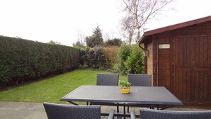 Une terrasse avec une table et des chaises, entourée d’une haie et d’un bâtiment en bois.