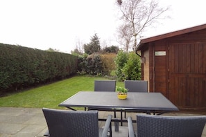 Eine Terrasse mit Tisch und Stühlen, umgeben von einer Hecke und einem Holzgebäude.