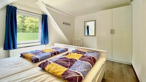 Une chambre avec un plafond en pente, une fenêtre avec des rideaux, un lit recouvert d’une couvre-lit à motifs et une armoire.