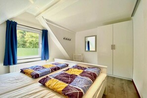 Ein Schlafzimmer mit schrägen Wänden, einem Fenster mit Vorhängen, einem Bett mit gemustertem Überwurf und einem Kleiderschrank.