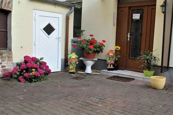 Una entrada de edificio con una puerta blanca, una maceta con flores rojas y una pequeña estatua en los escalones.