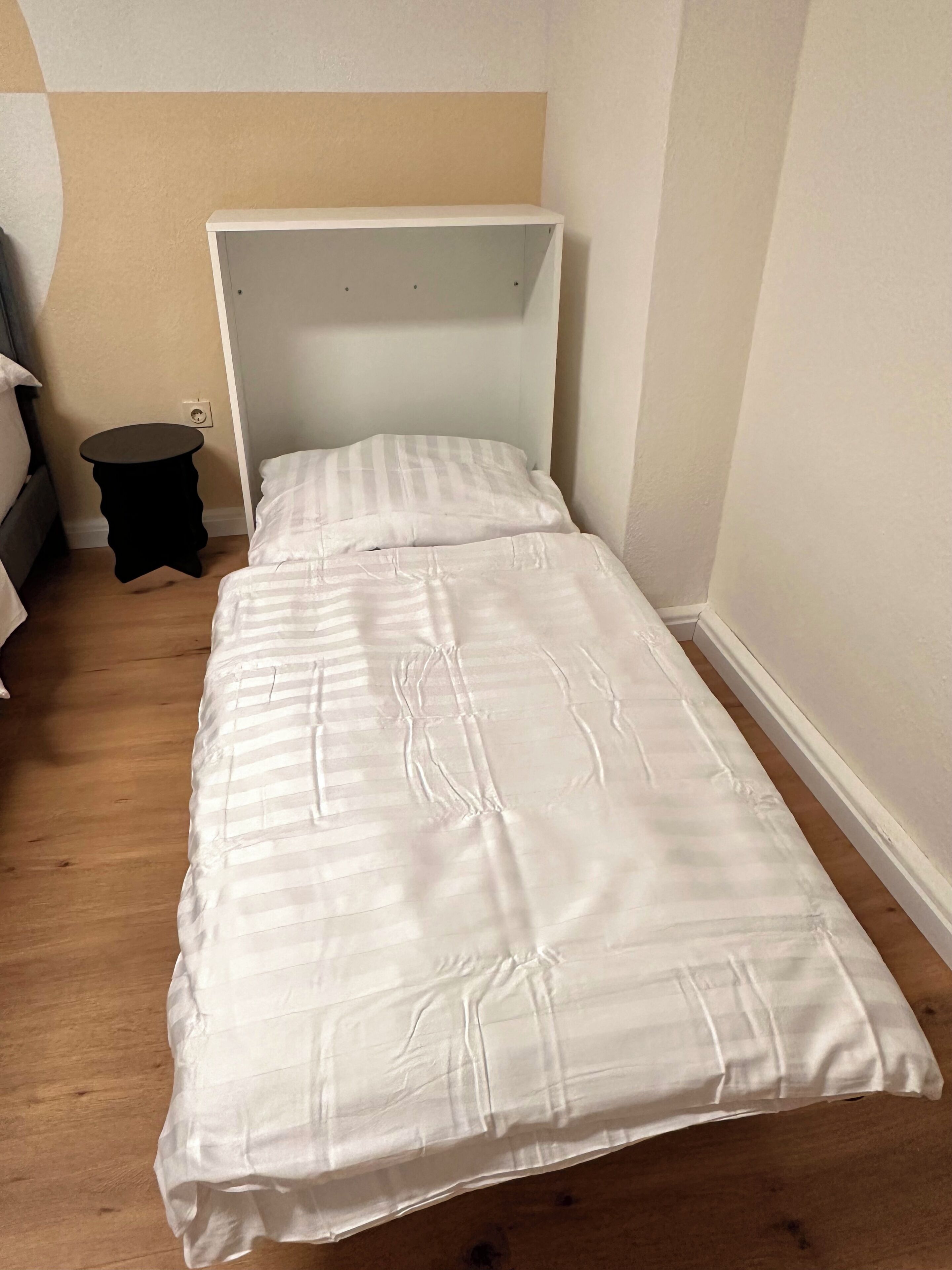Un lit simple avec une tête de lit blanche et une couvre-lit à rayures, dans une pièce au plancher en bois et où se trouve une petite table ronde.