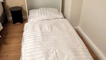 Un lit simple avec une tête de lit blanche et une couvre-lit à rayures, dans une pièce au plancher en bois et où se trouve une petite table ronde.
