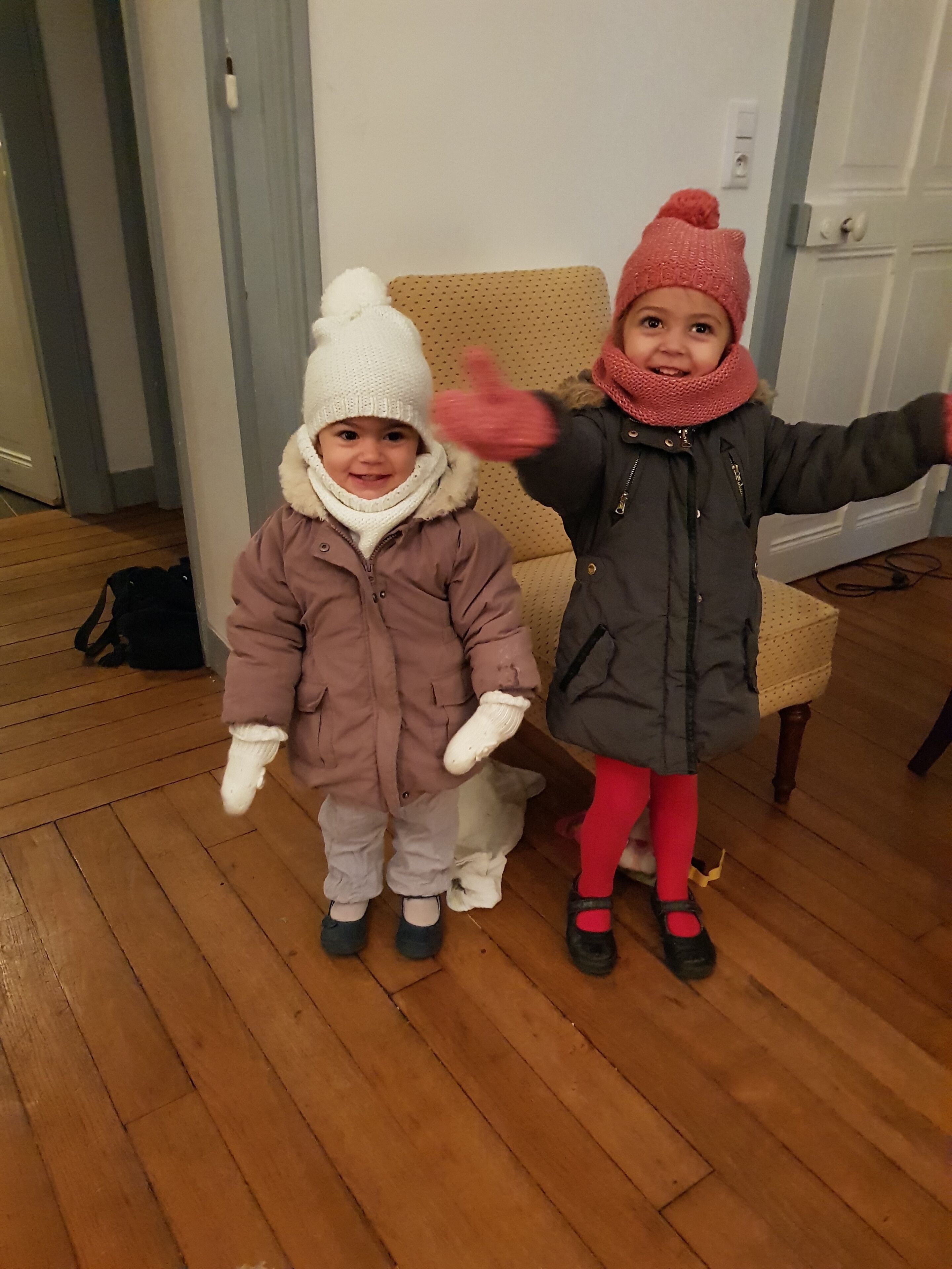 Zwei Kinder in Winterkleidung stehen in einem Raum mit Holzboden, einem Stuhl und einer Tür im Hintergrund.