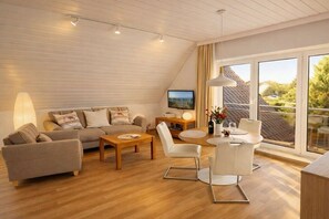 A modern living room with a sofa, coffee table, and chairs, a television, and a large window with a view. - Holiday apartment with 1 bedroom (Kellenhusen)