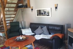 A living room with a black leather sofa, a green armchair, and a small orange table. There is a white furry throw blanket draped over the sofa. - |---former stable---| (Langelsheim)