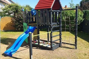 A playground with a slide, climbing net, and a red-roofed playhouse. - |---former stable---| (Langelsheim)