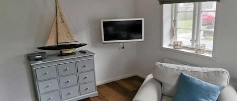 Una habitación con un modelo de velero, televisor, cómoda gris, sillón blanco y ventana con vistas al exterior.