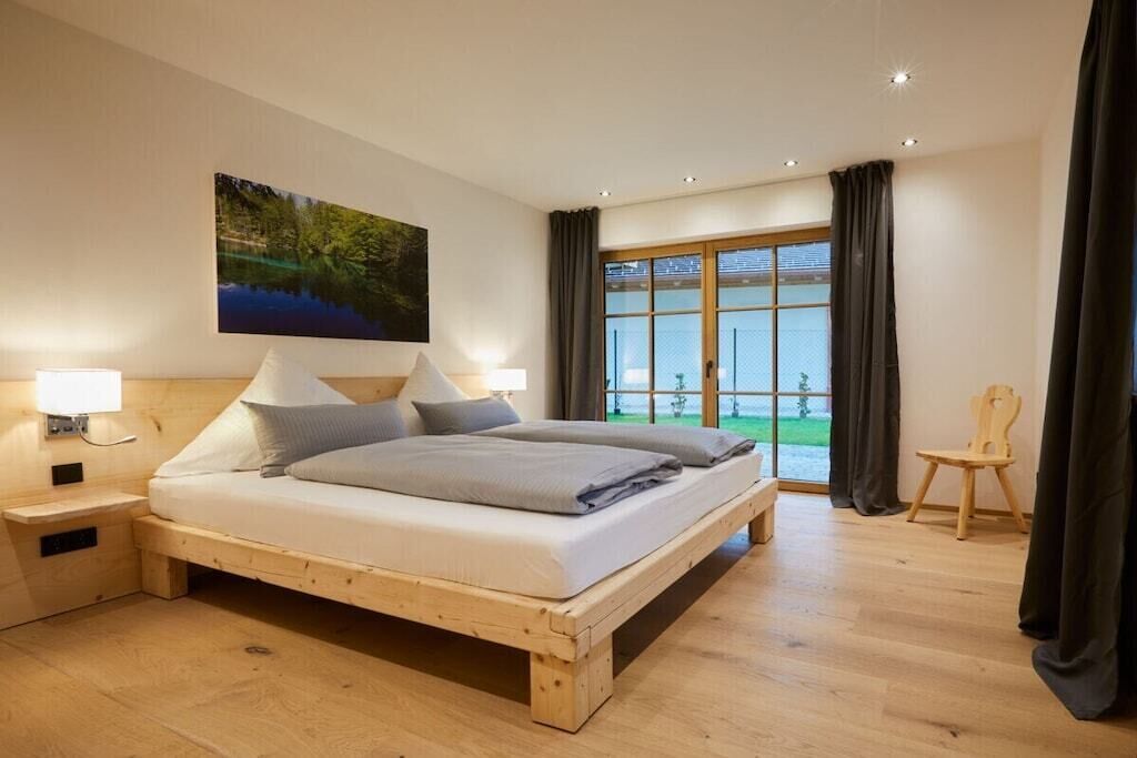 A bedroom with a wooden bed, a nightstand, a wall-mounted lamp, a window with curtains, and a view of greenery outside.