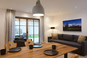 A modern living room with a sofa, dining table, and chairs. There is a large window with curtains, a painting on the wall, and a lamp. - WALDECK in the CHALET ZUGSPITZEREI (Grainau)