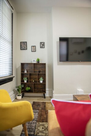 Ein Wohnzimmer mit an der Wand montiertem Fernseher, einer Holzregalanlage mit Dekoration und zwei Sesseln.