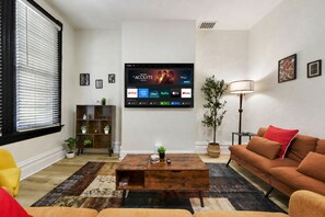 Ein Wohnzimmer mit einem Flachbildfernseher, der einen Film zeigt, einem braunen Sofa, einem gelben Sessel, einem Holztisch und einem Bücherregal mit Pflanzen.