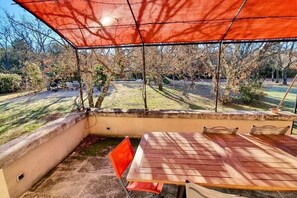 An outdoor patio with a table and chairs, a red canopy, and a view of a garden with trees and a lawn. - Charming country house with pool – 637 SAT (Saint Saturnin lès Apt)