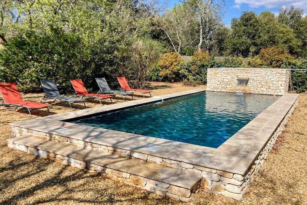 Piscina retangular com borda de pedra, rodeada por uma área gramada com espreguiçadeiras vermelhas e cinzas.
