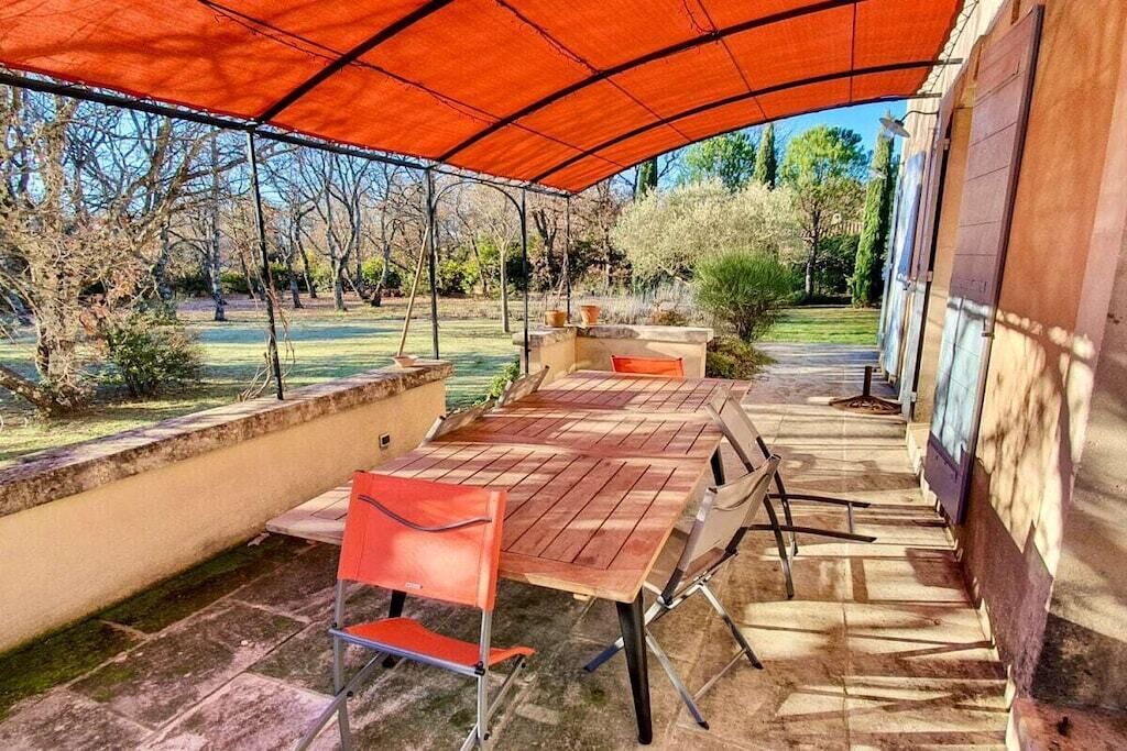 Un patio al aire libre con una mesa y sillas de madera, un paraguas rojo y vistas a un jardín.