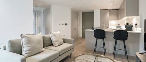 Een moderne woonkamer met een bank, twee stoelen, een glazen salontafel en een keukenhoek met een spoelbak en fornuis.