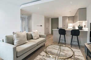 A modern living room with a sofa, two chairs, a glass coffee table, and a kitchen area with a sink and stove. - Paddington Green by Veeve (London)