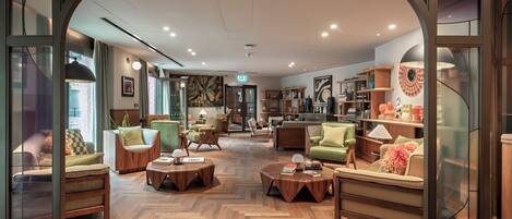A modern hotel lobby with a herringbone wood floor, multiple seating areas, and a central coffee table.