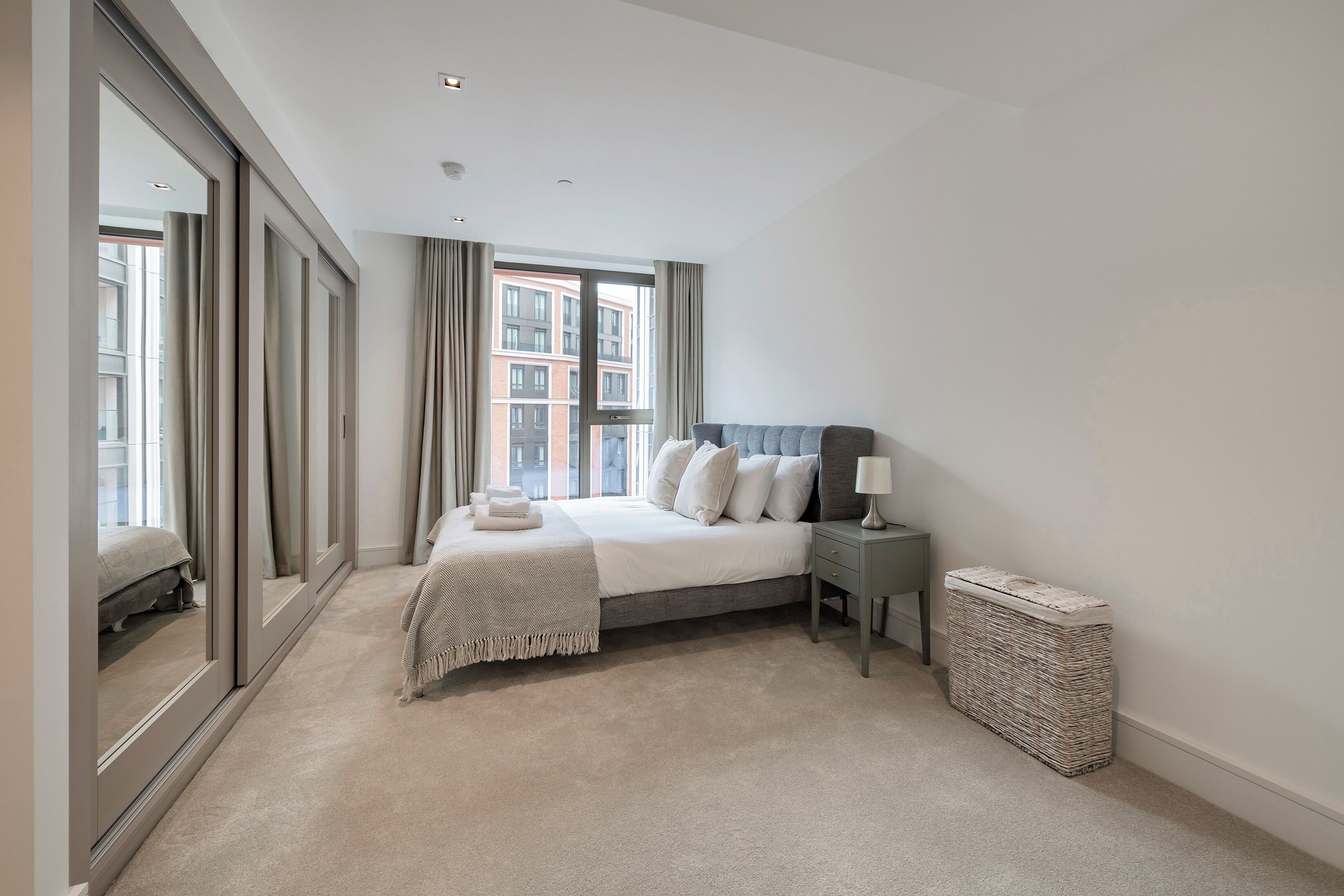 A bedroom with a large bed, a bedside table with a lamp, a small side table, a large mirror, and a window with curtains.