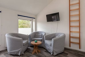A room with two grey armchairs, a small wooden table with a green object, a TV mounted on the wall, and a wooden ladder. - Willebrordstraat 13 Studio 3 (Westkapelle)