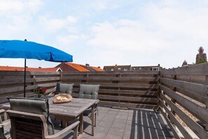A rooftop patio with a wooden table and chairs, a blue umbrella, and a view of buildings in the background. - Willebrordstraat 13 Studio 3 (Westkapelle)