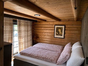 A wooden cabin room with a bed, plaid curtains, a framed picture on the wall, and a window with a view.