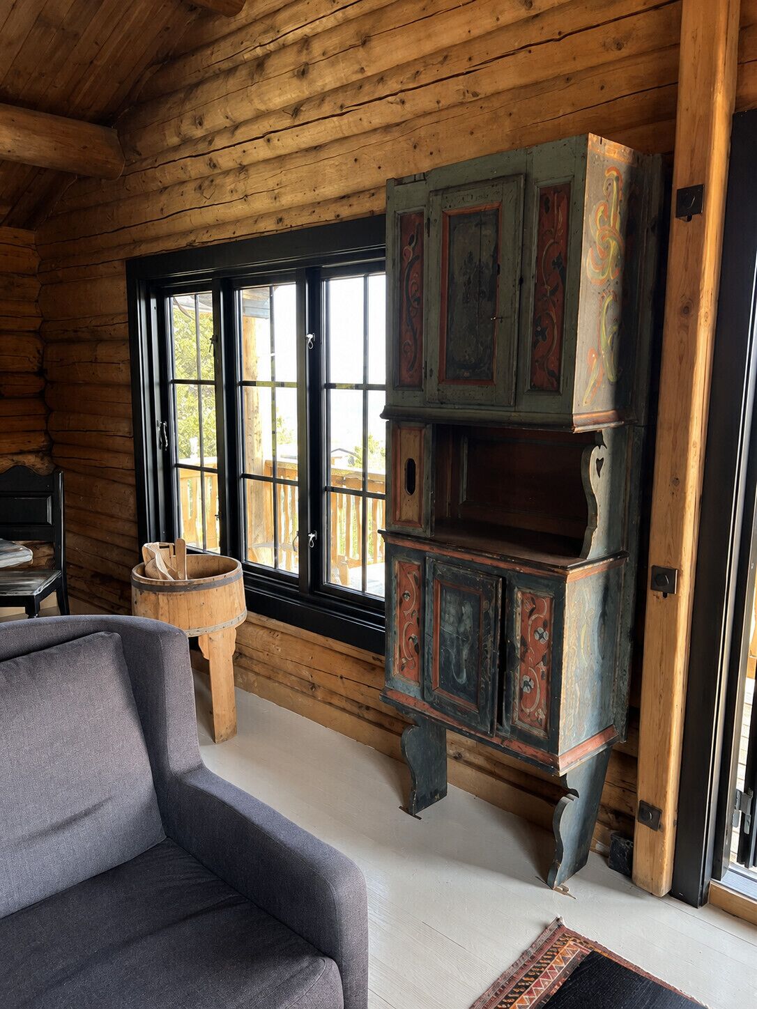 Interior de una cabaña de troncos con un sofá, un mueble de madera con detalles decorativos y un ventanal amplio.