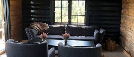 A log cabin interior with a dark-colored sofa, two armchairs, and a coffee table. There is a window with a view of trees, and a wooden ceiling with exposed beams.