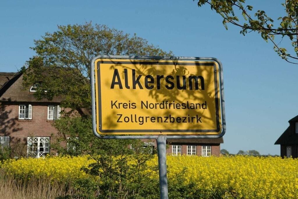 A yellow sign with the text 'Alkersum Kreis Nordfriesland Zollgrenzbezirk' in front of a building with a field of yellow flowers.