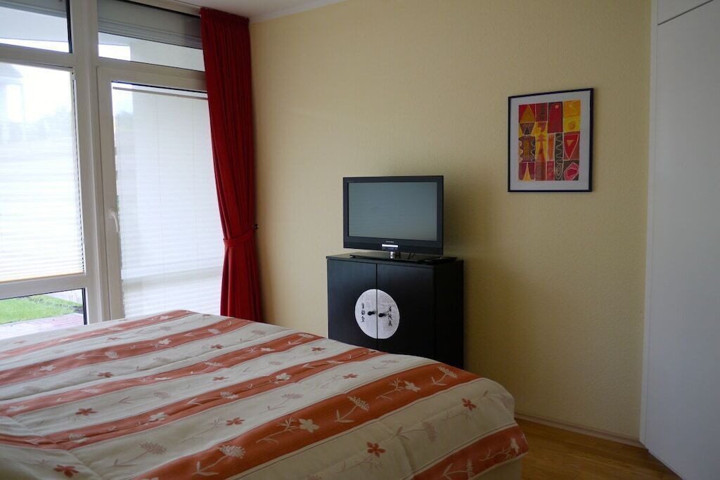 Ein Schlafzimmer mit einem Bett, einem Fernseher auf einer Kommode, einem Fenster mit Vorhängen und einem Wandbild.