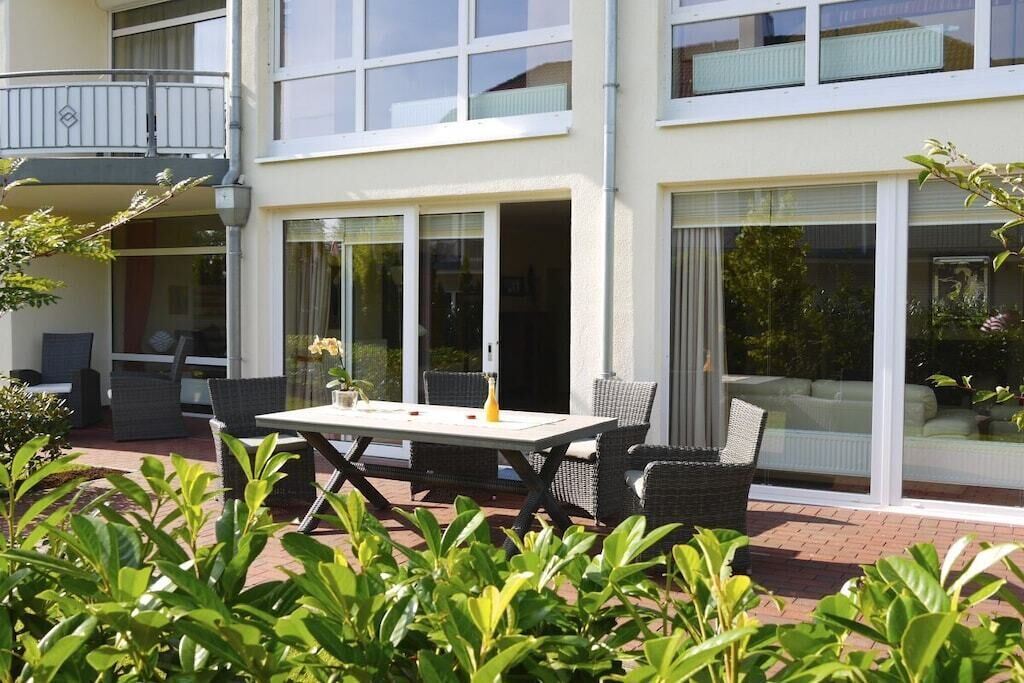 Un bâtiment moderne à deux étages, doté d’un balcon, d’un espace extérieur aménagé avec des meubles en osier et de grandes fenêtres.