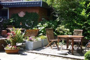 An outdoor patio area with wooden furniture, including a table and chairs, surrounded by potted plants and a brick wall. - House Rustica - Apartment 1 (Fehmarn)