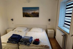 A bedroom with a bed, a window with blinds, a radiator, and a painting on the wall. - House Rustica - Apartment 1 (Fehmarn)