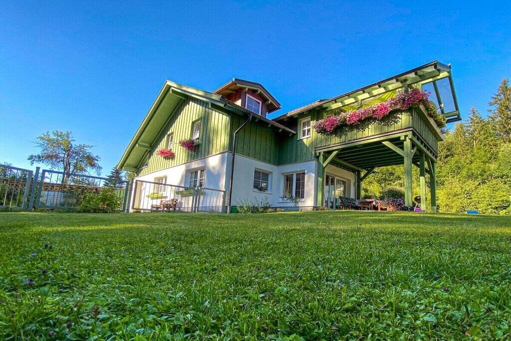 Una casa de dos pisos con fachada verde, un porche cubierto con plantas y un césped bien cuidado.