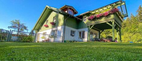 A two-story house with a green exterior, a covered porch with plants, and a well-maintained lawn.