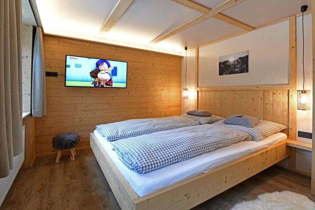 A wooden-paneled room with a bed, a television mounted on the wall, and a small stool.