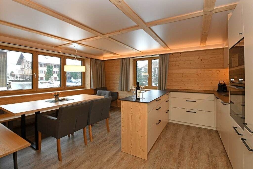 Una cocina moderna con armarios de madera, una zona de comedor con mesa y sillas, y grandes ventanales que ofrecen vistas al exterior.