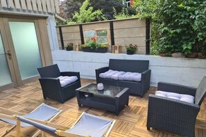 Eine Terrasse mit Korbmöbeln, einem Glastisch und Blick auf einen Garten mit Blumentöpfen.