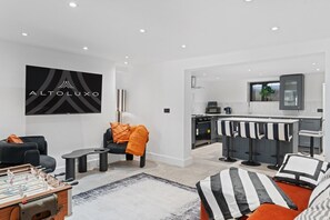A modern living room with a foosball table, a TV mounted on the wall, and a kitchen area with bar stools. - Large 5BR Home in Newport | Ideal for Work Crews | Sleeps 12 (Newport)