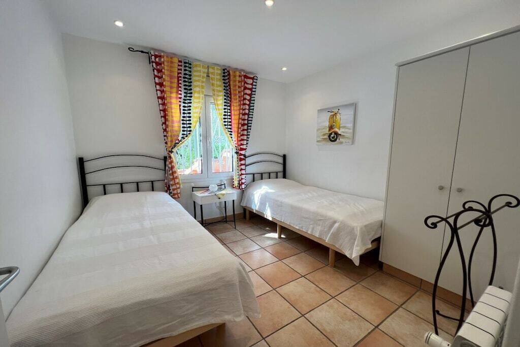 A bedroom with two single beds, a small bedside table, a window with colorful curtains, and a radiator.