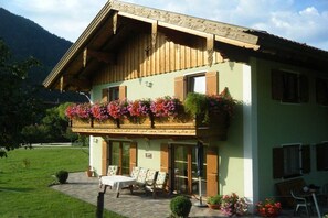 Un edificio de dos pisos con un balcón decorado con flores, un patio con mesa y sillas, y montañas al fondo.