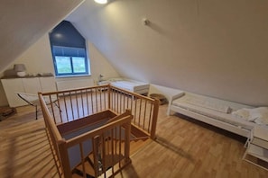 A small, sloped room with a wooden crib, a white sofa, and a window with blinds. - Holiday apartment with 1 bedroom (Friedrichskoog)