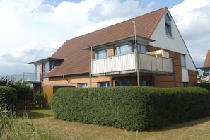 A two-story residential building with a sloped roof, a balcony on the second floor, and a well-maintained hedge in the front yard. - Holiday apartment with 1 bedroom (Friedrichskoog)