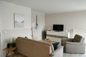 A modern living room with a sofa, armchair, and a television on a white cabinet. - Holiday apartment with 1 bedroom (Friedrichskoog)