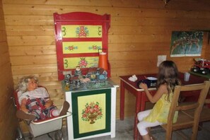 Interior d'una cabana de fusta amb una taula de menjador, cadires, un petitet frigorífic i una caseta de nines.