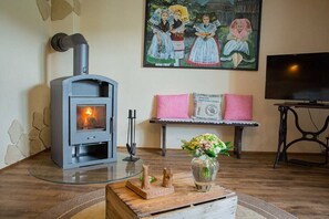 Ein Zimmer mit Kaminofen, einem Bild an der Wand, einer Bank mit karierten Kissen, einem Fernseher und einer Tischvase mit Blumen.
